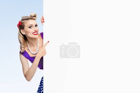 Foto de Mujer sonriente feliz en vestido de estilo pin-up, mostrando letrero en blanco con área de copyspace para el eslogan o mensaje de texto publicitario, sobre fondo azul. Modelo rubio caucásico en moda retro y concepto vintage. - Imagen libre de derechos