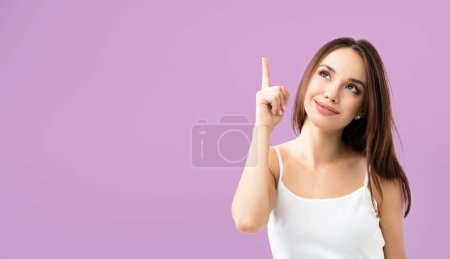 Photo for Smiling looking up young woman in white casual smart clothing, showing empty blank copyspace area for advertising text or slogan, over purple background - Royalty Free Image