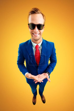 Téléchargez les photos : Et alors ? Portrait de jeune homme d'affaires drôle en lunettes de soleil, costume bleu et cravate rouge, vue de dessus angle, sur fond jaune-orange. Concept d'entreprise. - en image libre de droit