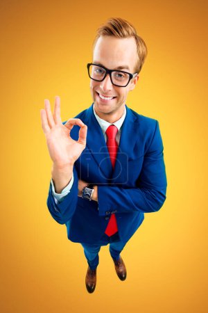 Téléchargez les photos : OK! Portrait of funny happy businessman in glasses, blue suit and red tie, showing okay gesture or zero, top angle view shot, over yellow-orange background. Business concept. - en image libre de droit