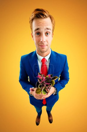 Porträt eines lustigen jungen Geschäftsmannes mit Brille, blauem Anzug und roter Krawatte, Blumentopf mit Blumen in der Hand, Aufnahme aus der Vogelperspektive, vor gelb-orangefarbenem Hintergrund. Geschäftskonzept.