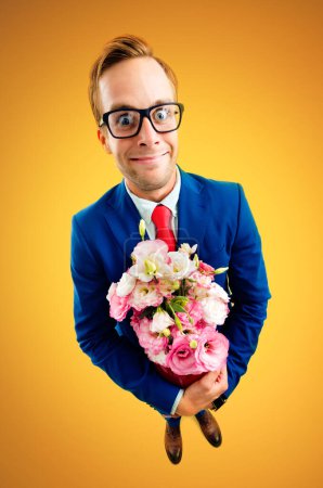 Porträt eines lustigen jungen Geschäftsmannes mit Brille, blauem Anzug und roter Krawatte, Blumenstrauß in der Hand, Aufnahme aus der Vogelperspektive, vor gelb-orangefarbenem Hintergrund. Geschäftskonzept.