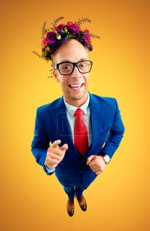 Téléchargez les photos : Portrait of funny happy businessman in glasses, confident blue suit, red tie and wreath of wildflowers on his head, pointing on you, top angle view shot, over yellow-orange background. - en image libre de droit
