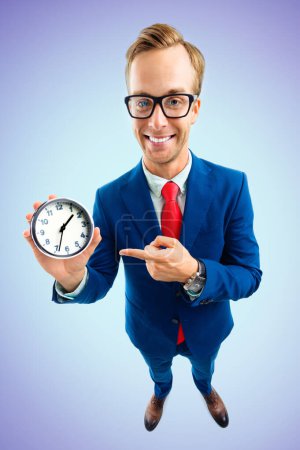 Portrait of funny happy businessman in glasses showing clock, confident suit and red tie, top angle view shot, on blue background. Business and time concept.