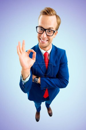 Foto de ¡Bien! Retrato de empresario feliz divertido en gafas, traje seguro y corbata roja, mostrando gesto bien o cero, vista de ángulo superior, sobre fondo azul. Concepto empresarial. - Imagen libre de derechos