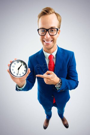 Portrait of funny happy businessman in glasses showing clock, blue suit and red tie, top angle view shot, over grey background. Business and time concept.