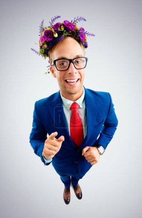 Téléchargez les photos : Portrait de drôle homme d'affaires heureux dans des lunettes, costume bleu confiant, cravate rouge et couronne de fleurs sauvages sur sa tête, pointant sur vous, vue de dessus angle, sur fond gris. - en image libre de droit