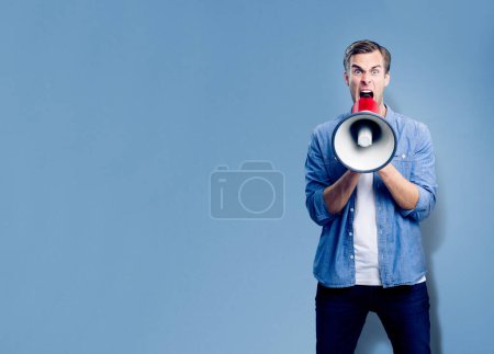 Homme criant à travers le mégaphone, avec espace de copie vide pour slogan, publicité ou message texte, sur fond bleu. Modèle masculin caucasien habillement décontracté intelligent faisant annonce, concept studio.