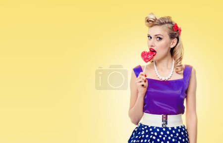 Photo for Woman eating heart shape lollipop dressed in pinup style dress, with copyspace area for slogan or advertising text message, on yellow background. Caucasian blond model in retro fashion and vintage concept. - Royalty Free Image