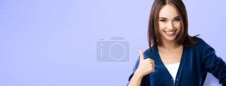 Photo for Smiling beautiful young woman in casual smart blue clothing, showing thumbs up gesture, over violet background, with copyspace for slogan, advertising or text message. Banner composition. - Royalty Free Image