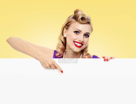 Photo for Smiling blond woman in pin-up style dress, showing blank signboard with copyspace, on yellow background. Caucasian blond model posing in retro fashion and vintage concept studio shoot. - Royalty Free Image