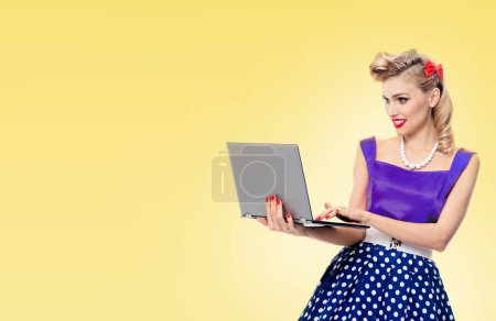 Téléchargez les photos : Femme tenant un ordinateur portable, vêtue d'une robe de style pin-up à pois, avec espace de copie pour slogan ou message publicitaire, sur fond jaune. Modèle blond caucasien en mode rétro et concept vintage. - en image libre de droit