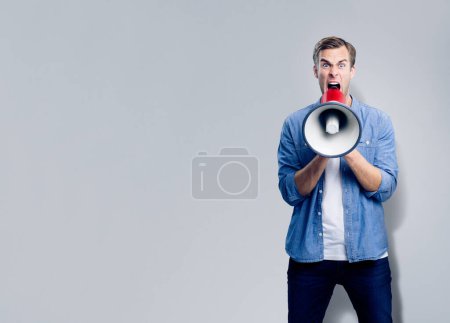 Man shouting through megaphone, with empty copyspace area for slogan, advertising or text message, over grey background. Caucasian male model in blue smart casual clothing making announcement, studio concept.