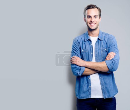 Porträt eines glücklich lächelnden Mannes, der in verschränkten Armen vor grauem Hintergrund posiert, mit leerem Kopierraum für Slogans, Werbung oder Textnachrichten. Kaukasisches männliches Modell in schicker Freizeitkleidung, Studioaufnahme.