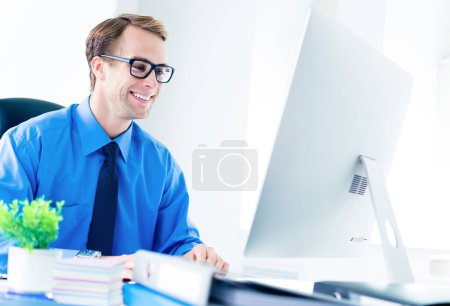 Foto de Feliz joven empresario sonriente en ropa segura, camisa azul, corbata y gafas, trabajando con computadora de escritorio en la oficina. Éxito en el concepto de empresa, empleo y educación. - Imagen libre de derechos