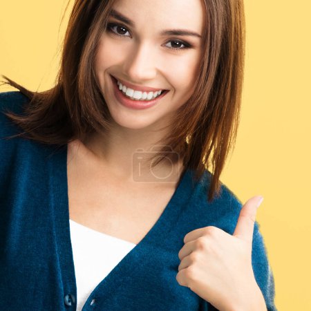 Téléchargez les photos : Jeune femme heureuse en vêtements bleus intelligents décontractés, montrant les pouces vers le haut geste, sur fond jaune - en image libre de droit
