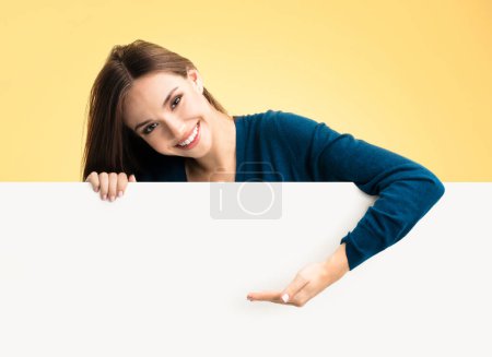 Téléchargez les photos : Femme heureuse dans les vêtements intelligents décontractés bleus, montrant le panneau blanc vide avec la zone d'espace de copie pour une certaine publicité, texte ou slogan, sur fond jaune - en image libre de droit