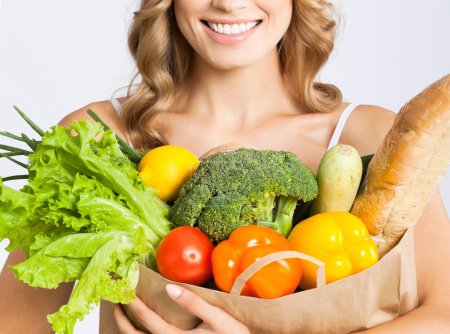 Téléchargez les photos : Femme souriante tenant sac d'épicerie avec de la nourriture crue végétarienne saine, sur fond gris - en image libre de droit