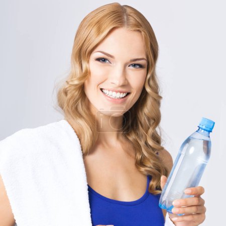 Photo for Happy smiling young attractive blond woman in fitness wear with bottle of water and towel, against grey background - Royalty Free Image