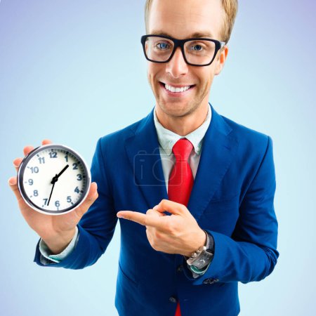 Funny happy businessman in glasses pointing at clock, confident suit and red tie, on blue background. Business and time concept.