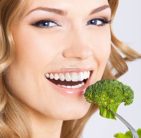 Téléchargez les photos : Portrait de jeune femme souriante heureuse mangeant du brocoli, sur fond gris - en image libre de droit