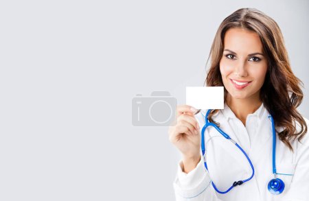 Photo for Portrait of happy smiling young female doctor showing blank business card or invitation, with copy space for some text, advertising or slogan, against grey background - Royalty Free Image