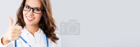 Happy smiling female doctor with thumbs up gesture, with copy space for some text, advertising or slogan, against grey background. Banner composition.