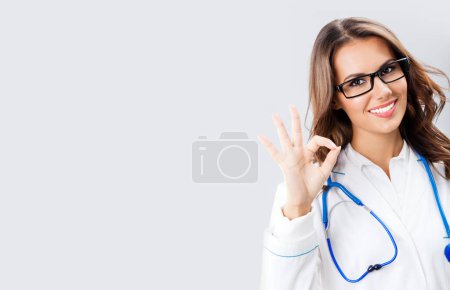 Happy smiling young female doctor showing okay gesture, with copy space for some text, advertising or slogan, against grey background