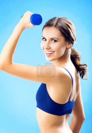 Photo for Portrait of young woman in fitness wear exercising with dumbbell, over blue background - Royalty Free Image