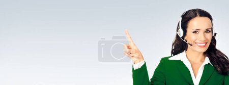 Foto de Feliz operador de teléfono de atención al cliente sonriente en traje verde, mostrando algo o área de espacio de copia para algún texto, publicidad o eslogan, sobre fondo gris - Imagen libre de derechos