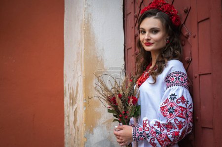 Téléchargez les photos : Portrait d'une belle fille slave aux longs cheveux blonds et aux yeux bleus avec une couronne de fleurs dans une robe brodée blanche et rouge avec un bouquet. Vêtements traditionnels de la région ukrainienne - en image libre de droit