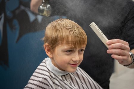 Photo for Happy cute fair-haired preschool boy getting a haircut. Children's hairdresser with scissors and comb cuts a little boy's hair in a room with a loft interior - Royalty Free Image