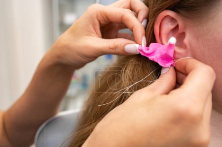 Téléchargez les photos : Un médecin remplit le conduit auditif d'un patient avec un matériau en silicone, rose, fabriqué à l'aide d'une seringue pour fabriquer des bouchons d'oreille dans une clinique auditive. Production d'inserts sur commande - en image libre de droit