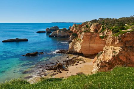 Foto de Playa solitaria cerca del pueblo de Alvor en Portugal. Hay un montón de rocas de piedra caliza en el agua y hierba verde en primer plano. - Imagen libre de derechos