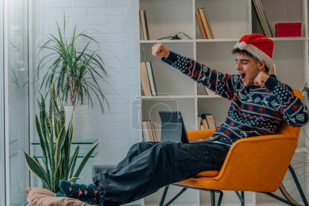 Téléchargez les photos : Jeune homme à Noël à la maison célébrant excité regardant ordinateur portable ou tablette - en image libre de droit