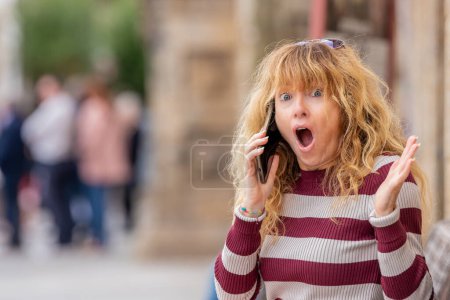 Téléchargez les photos : Femme d'âge moyen parlant dans la rue avec un téléphone portable ou cellulaire et expression de surprise - en image libre de droit