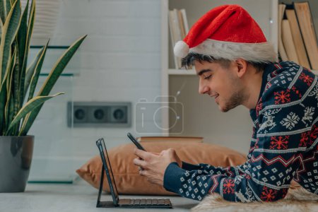 Téléchargez les photos : Jeune homme avec ordinateur et téléphone portable à la maison à Noël avec chapeau Père Noël - en image libre de droit