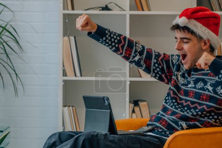 Photo for Young man at christmas at home celebrating excited looking at laptop or tablet - Royalty Free Image