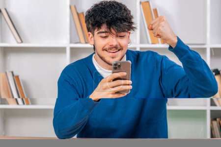 Téléchargez les photos : Jeune étudiant au bureau excité en regardant smartphone - en image libre de droit