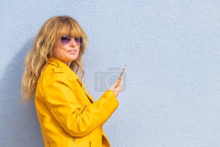 Téléchargez les photos : Femme d'âge moyen avec téléphone portable isolé sur le mur de la rue avec copie-espace - en image libre de droit