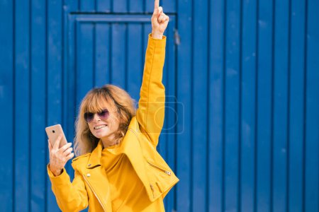 mujer de mediana edad feliz con teléfono móvil sobre fondo azul en la calle