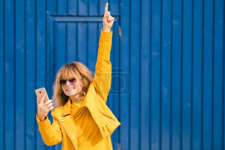 Foto de Mujer de mediana edad feliz con teléfono móvil sobre fondo azul en la calle - Imagen libre de derechos