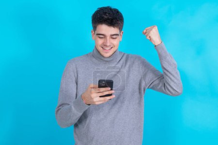 Foto de Joven celebrando emocionado con el teléfono móvil o teléfono inteligente aislado sobre fondo azul - Imagen libre de derechos