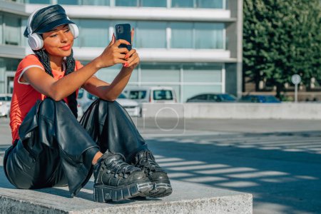Téléchargez les photos : Fille assise dans la rue avec écouteurs et téléphone mobile - en image libre de droit