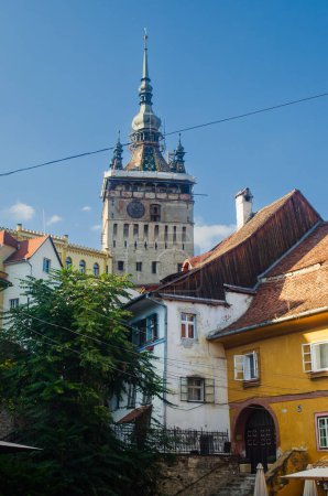 Foto de Antigua torre medieval histórica del reloj rodeada de casas pintadas de amarillo y blanco, edificios con techos de azulejos y chimeneas de ladrillo en Sighisoara Rumania - Imagen libre de derechos