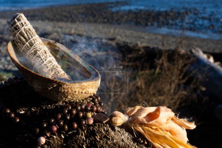 Photo for A burning smudge stick with abalone shell and mala beads on a driftwood stump with the receding tide in the background. - Royalty Free Image