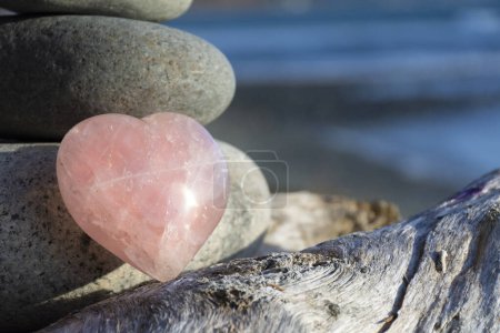Eine Nahaufnahme eines schönen herzförmigen Rosenquarzes Kristall lehnt an gestapelten Zen-Steinen mit dem blauen Ozean im Hintergrund. 