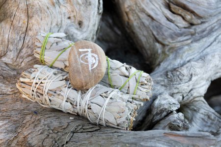 A close up image of two white sage smudge sticks and smooth stone with a healing symbol resting on old weathered driftwood. 