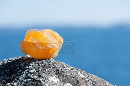 Foto de Una imagen de cerca de un cristal de calcita naranja sin pulir descansando sobre una roca cubierta de liquen con un fondo azul océano. - Imagen libre de derechos
