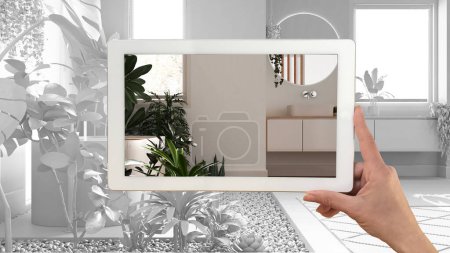 Augmented reality concept. Hand holding tablet with AR application used to simulate furniture and design products in total white unfinished background, urban jungle bathroom